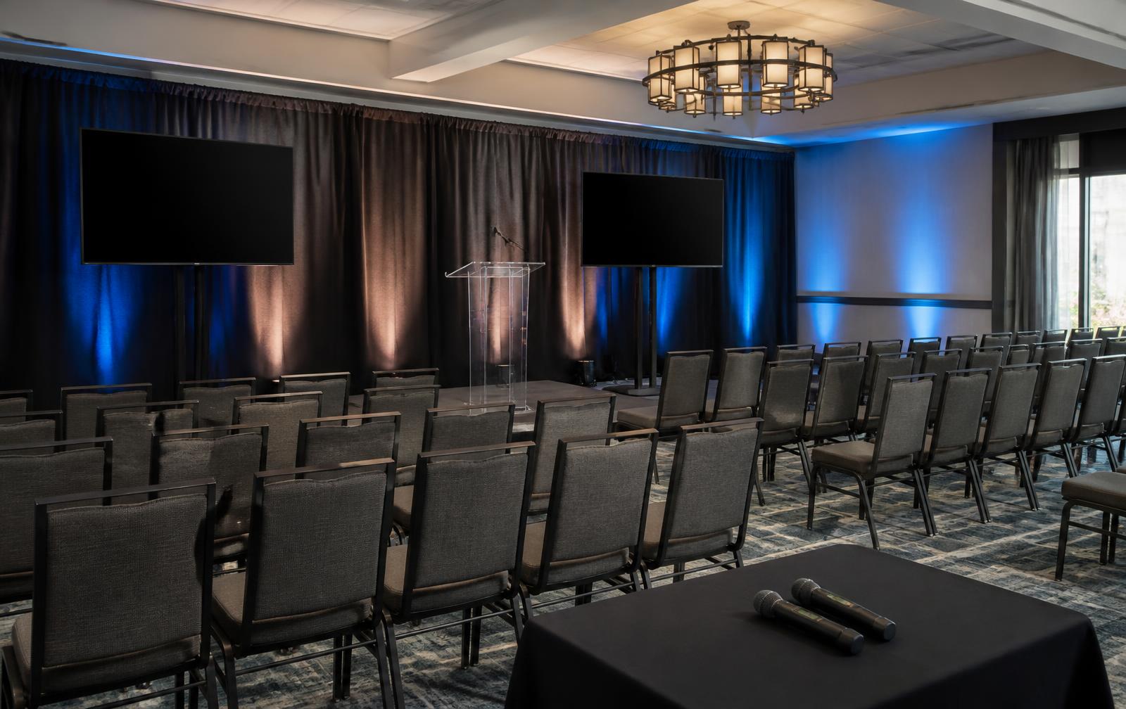 Intimate town hall setup with two displays, a clear lectern, charcoal draping, uplights and wireless microphones