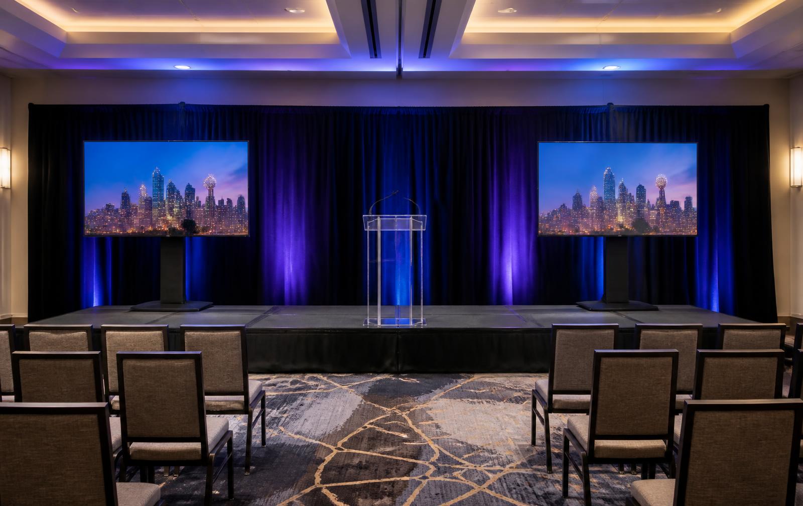 Small corporate meeting stage with two large TVs, a clear acrylic podium, black draping and blue-violet uplighting